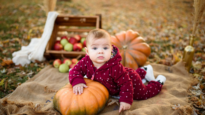 A Thanksgiving photo of a newborn in a onesie, surrounded by tiny pumpkins, leaves, and fall decor.