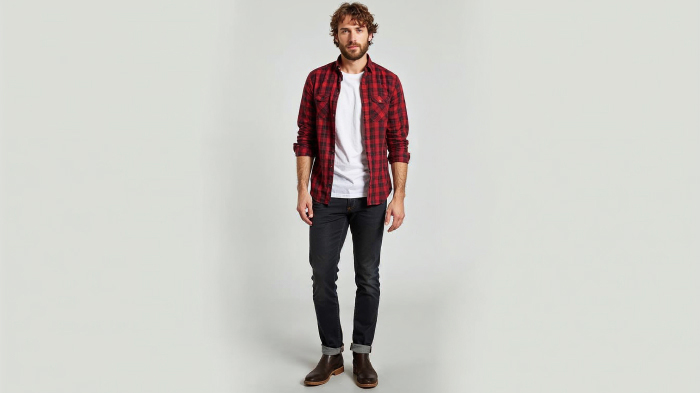 A photo of a man in a red plaid button-up shirt with a plain white t-shirt underneath, dark jeans, and casual boots.