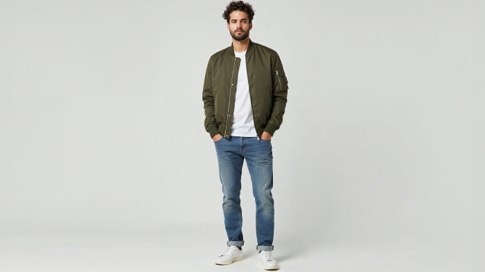 A photo of a man in an olive bomber jacket with a plain t-shirt, slim-fit jeans, and white sneakers.